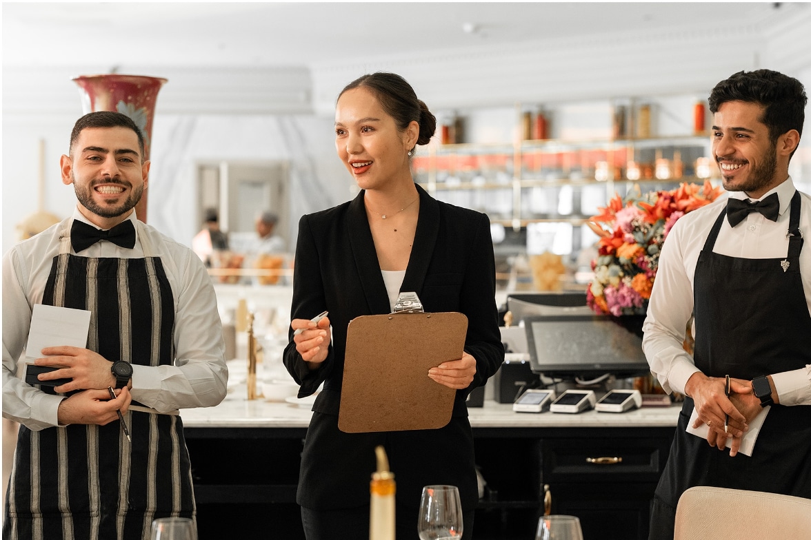 Three restaurant staff members, two men and one woman, smiling and interacting in a luxurious dining setting with a modern open kitchen in the background.