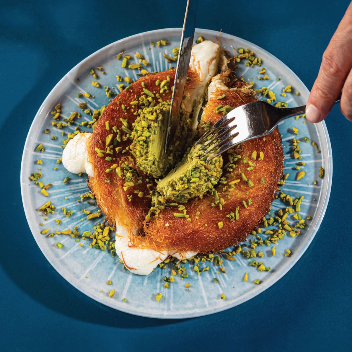 Close-up of a gourmet dessert with pistachio and cream on a blue plate, being eaten with a fork