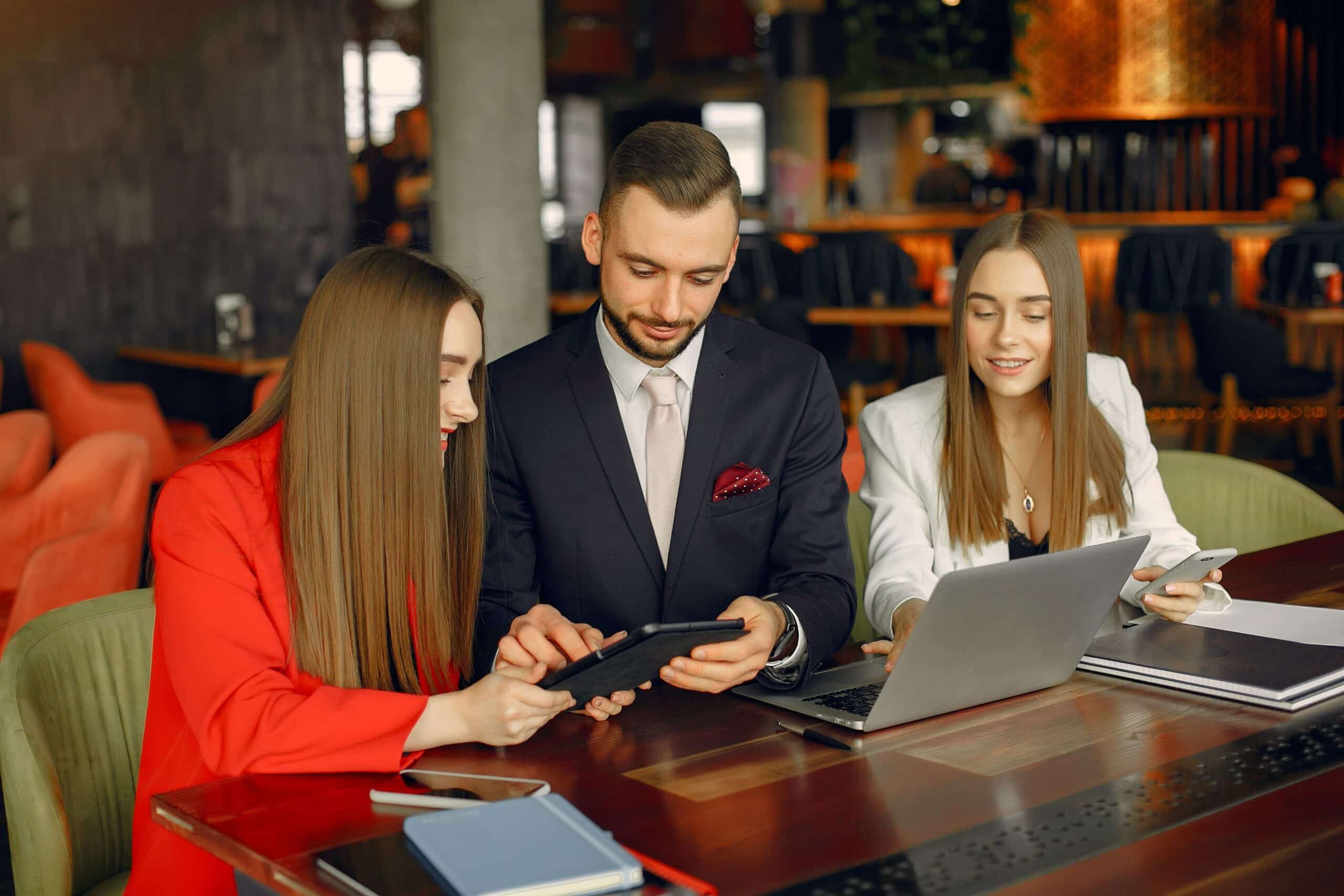 hree business professionals, two women and one man, in a modern office setting collaborating on a project. They are seated at a table, engaged with a laptop and tablet, discussing and smiling as they work together.