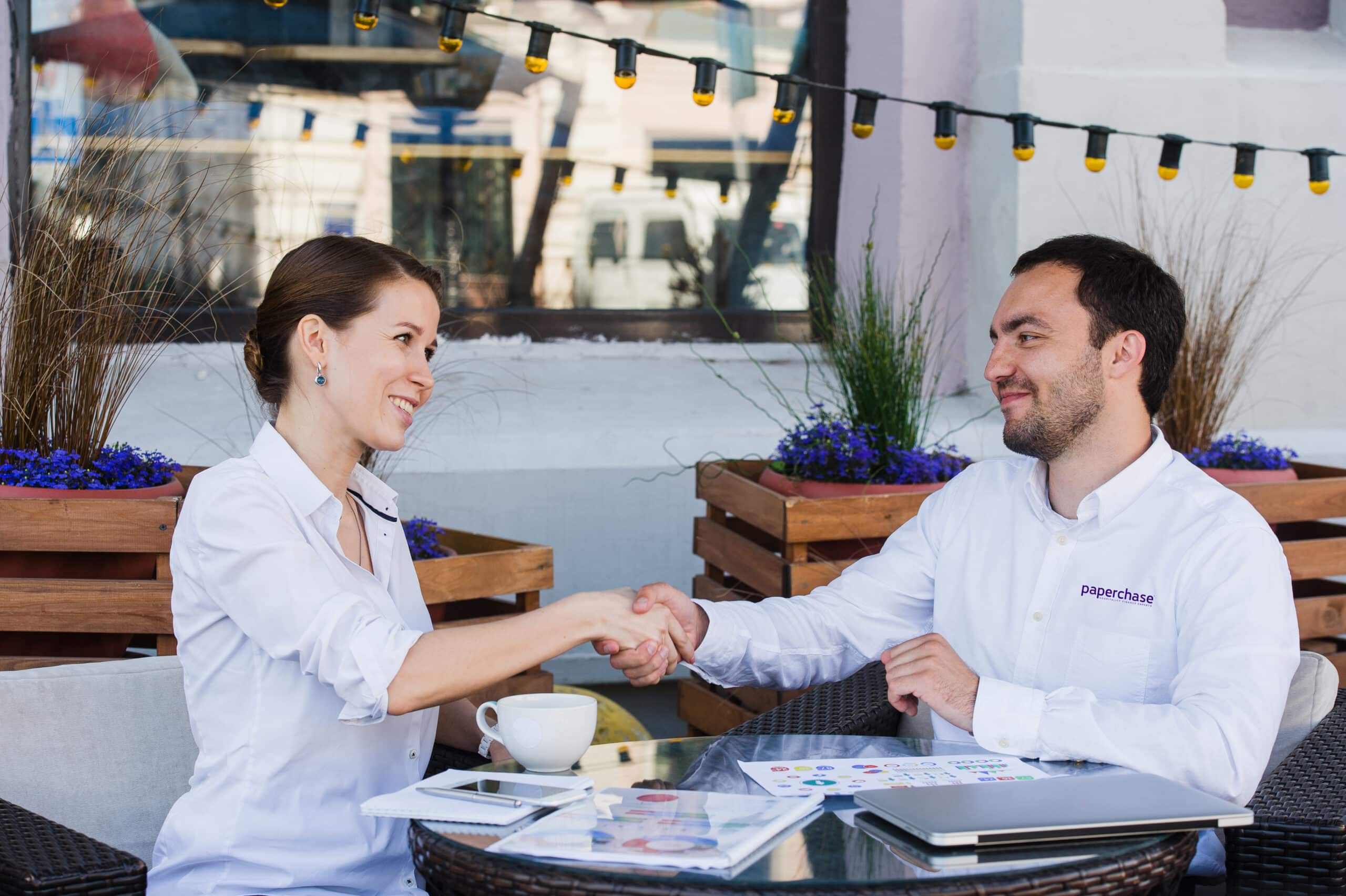 Two business professionals shaking hands in an outdoor cafe setting, smiling and engaged in a positive discussion, with documents and a laptop on the table.