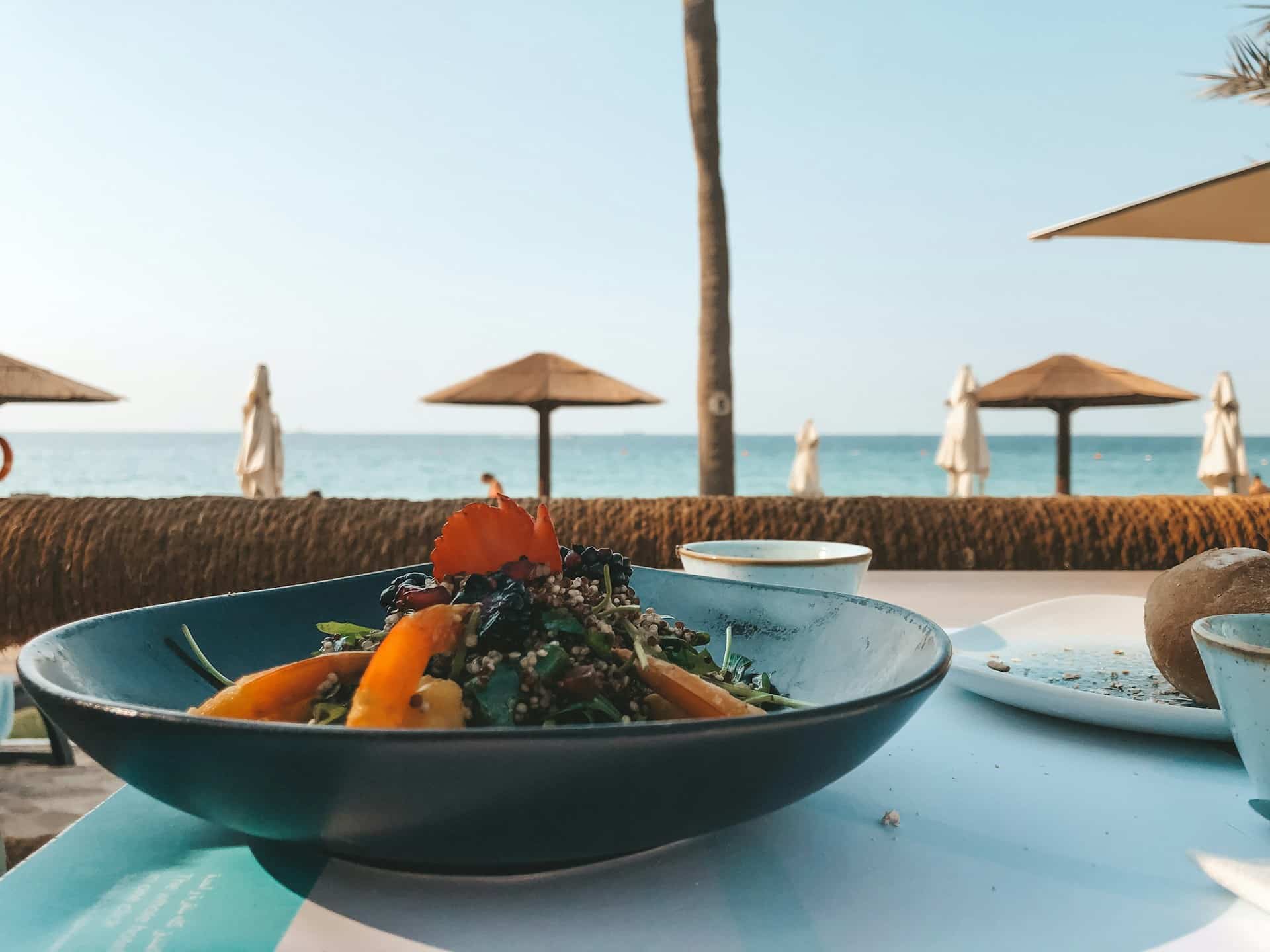 Healthy food served in a blue bowl on a beachside restaurant table with the ocean in the background and straw umbrellas on the sand