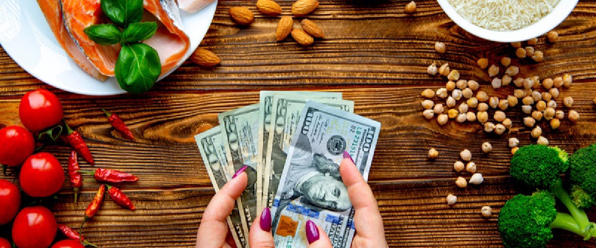 Hands holding dollar bills surrounded by fresh ingredients like vegetables, nuts, and fish, symbolizing recipe costing and food expenses