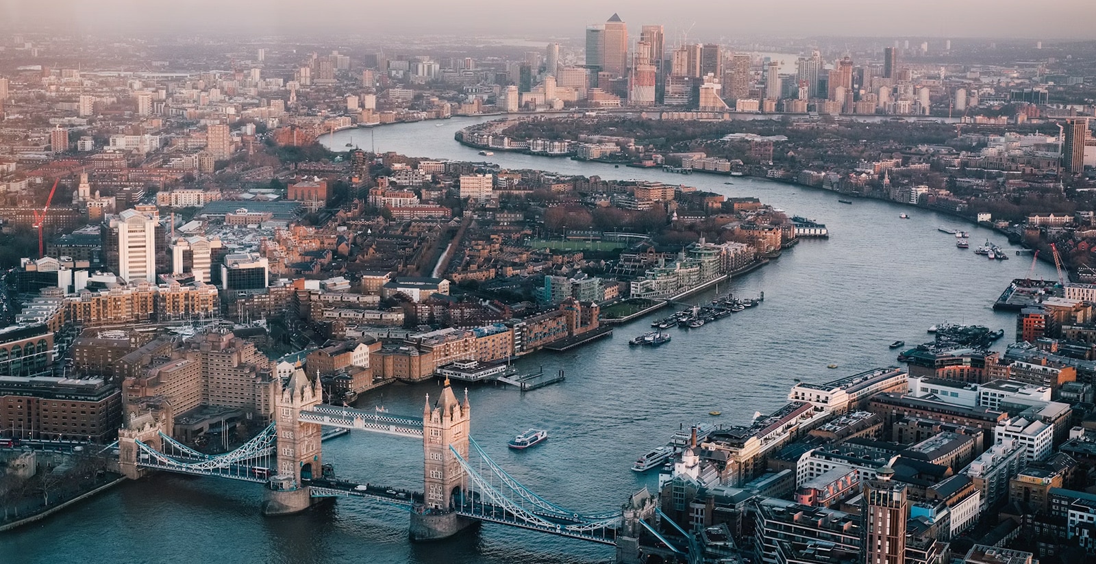 Aerial view of London cityscape with Tower Bridge spanning over the River Thames, showcasing surrounding modern and historical architecture under a hazy sky