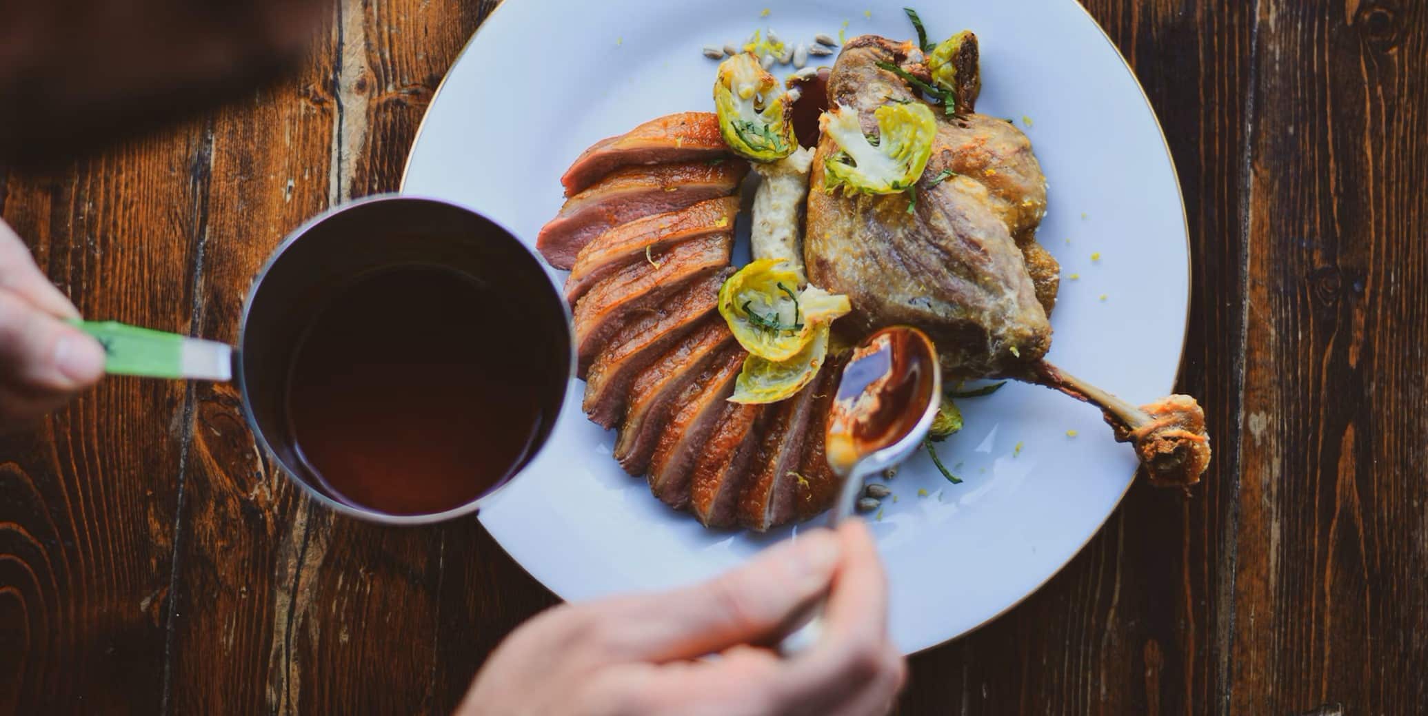A gourmet dish featuring sliced duck breast arranged in a fan shape on a white plate, accompanied by a crispy duck leg, garnished with roasted Brussels sprouts and served with a sauce. A person is pouring additional sauce from a small saucepan onto the plate. The background is a rustic wooden table.